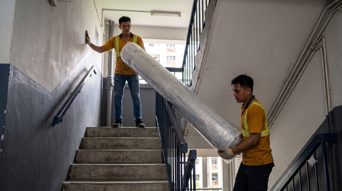 Two movers in hi-vis amber shirts carrying a rolled mattress down an HDB staircase
